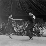 Coppia in abito da sera e completo esegue una salsa danza, con pubblico seduto sullo sfondo.