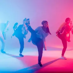 Un gruppo di ballerini esegue una coreografia hip hop con calci in aria, illuminati da luci blu e rosa.
