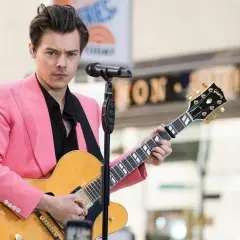 Harry Styles con giacca rosa suona la chitarra, ispirando tante canzoni da suonare con la chitarra.