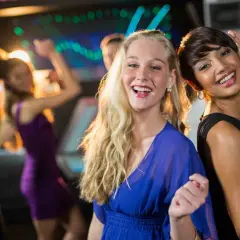 Due amiche sorridono mentre si divertono a ballare latino americano in discoteca, con luci colorate e altre persone che ballano sullo sfondo.