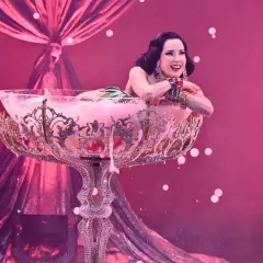 Attrice in un bicchiere gigante, un'esibizione di burlesque dance con schiuma rosa e drappeggi di seta.