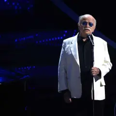 Un uomo con occhiali da sole blu canta sul palco, con un pianoforte a coda scuro e luci blu sullo sfondo. Sembra che sia lui chi ha scritto il cielo in una stanza.