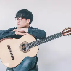 Giovane con occhiali appoggiato a una chitarra classica, pensieroso. Quanto tempo ci vuole per suonare la chitarra? Dipende dalla passione.