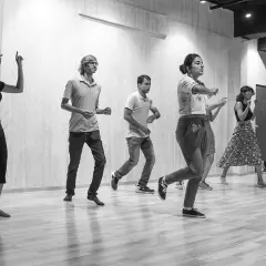 Un gruppo di persone impara come si balla lo swing in una sala prove. Movimenti energici e sorrisi contagiosi.