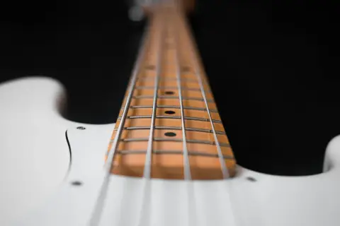 Primo piano del manico di un basso elettrico bianco, con le corde metalliche che corrono lungo la tastiera in legno chiaro.