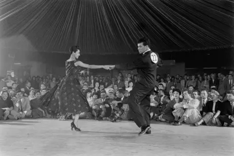 Coppia in abito da sera e completo esegue una salsa danza, con pubblico seduto sullo sfondo.