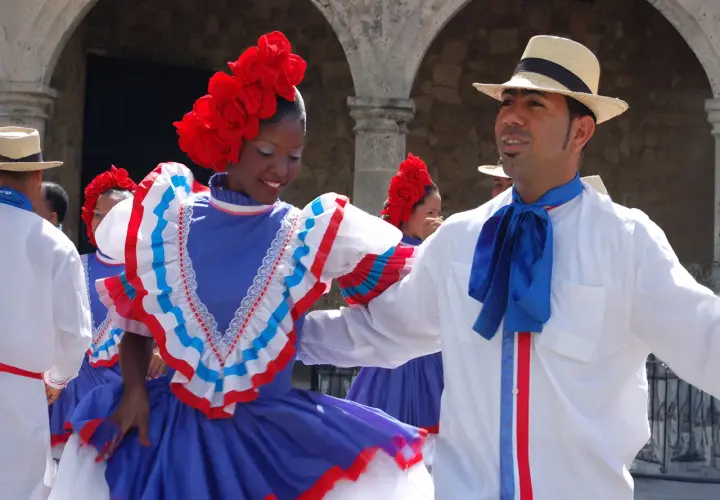 Coppia in abiti tradizionali, lei con fiori rossi sui capelli e un vestito blu con balze, lui con cappello di paglia e fiocco blu, pronti per un vivace ballo di salsa e merengue.