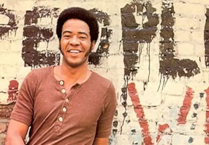 Bill Withers sorride davanti a un muro con graffiti. Anche se "ain't no sunshine", il suo sorriso illumina la scena.