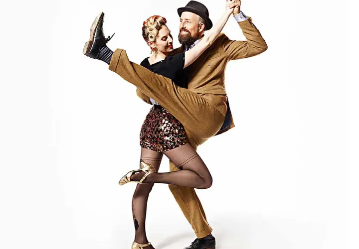 A man in a tan suit dips a woman in a sequined skirt during a Lindy Hop dance.