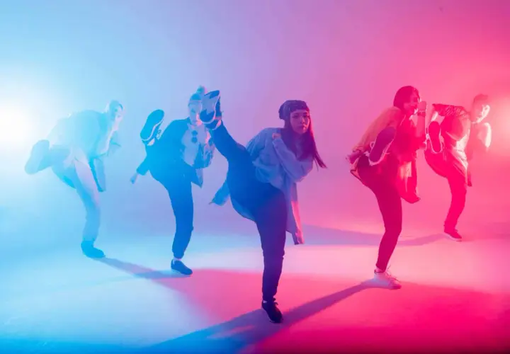 Un gruppo di ballerini esegue una coreografia hip hop con calci in aria, illuminati da luci blu e rosa.