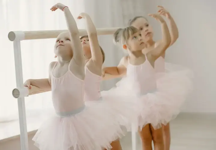 Bambine in tutù rosa praticano la danza propedeutica alla sbarra, con braccia alzate e sguardi concentrati.