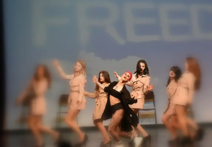 Un gruppo di ballerine in trench esegue una coreografia energica, con un'artista in primo piano che canta. Le canzoni di danza moderna creano un'atmosfera vibrante.