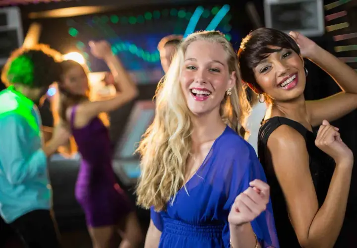 Due amiche sorridono mentre si divertono a ballare latino americano in discoteca, con luci colorate e altre persone che ballano sullo sfondo.