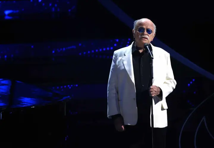 Un uomo con occhiali da sole blu canta sul palco, con un pianoforte a coda scuro e luci blu sullo sfondo. Sembra che sia lui chi ha scritto il cielo in una stanza.