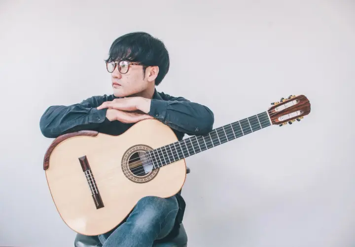 Giovane con occhiali appoggiato a una chitarra classica, pensieroso. Quanto tempo ci vuole per suonare la chitarra? Dipende dalla passione.