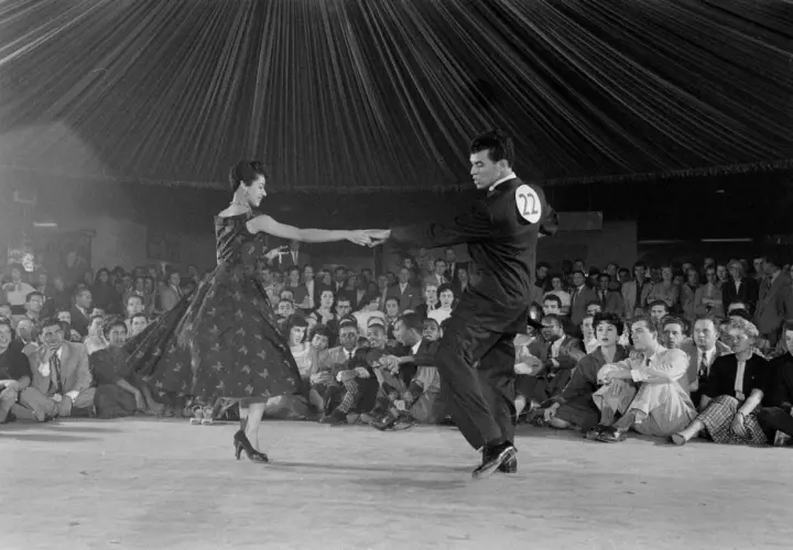 Coppia in abito da sera e completo esegue una salsa danza, con pubblico seduto sullo sfondo.