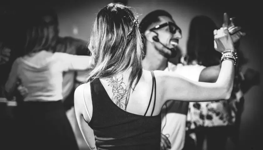 Coppie danzano in un corso di ballo latino americano. La donna in primo piano ha un tatuaggio sulla schiena.