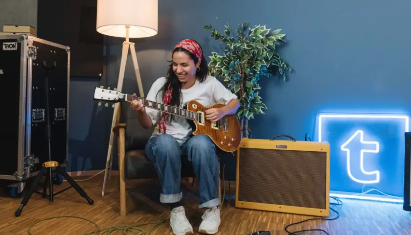 Donna sorridente suona la chitarra, con un'insegna al neon blu sullo sfondo che ricorda le "blue canzoni famose".