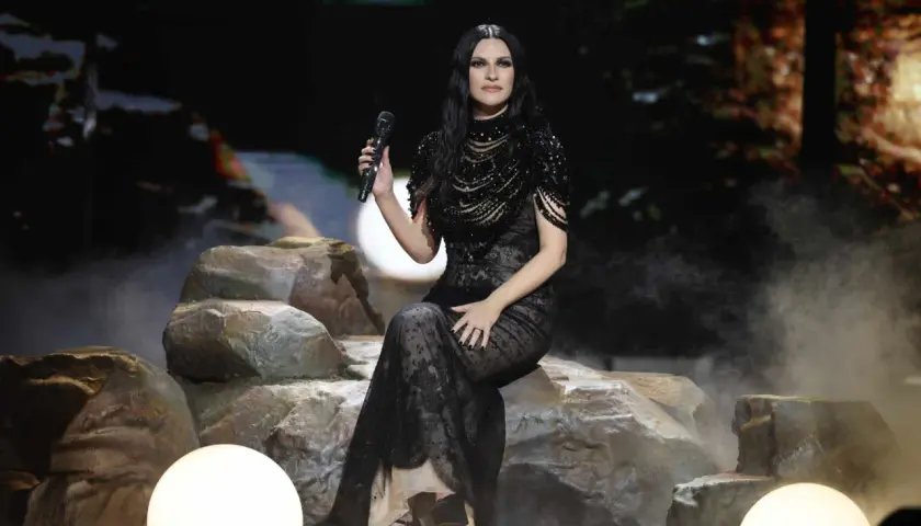 Lei di Laura Pausini, seduta su rocce con un microfono in mano, indossa un abito nero scintillante e trasparente, immersa in un'atmosfera fumosa.