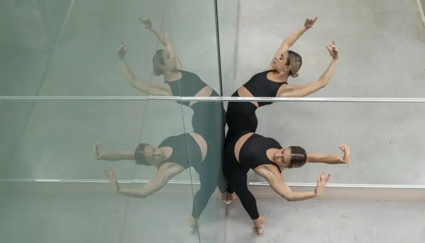 Due ballerine in abiti neri eseguono movimenti fluidi, riflesse su una parete specchiata. Un'immagine che evoca la grazia dei corsi di danza moderna.