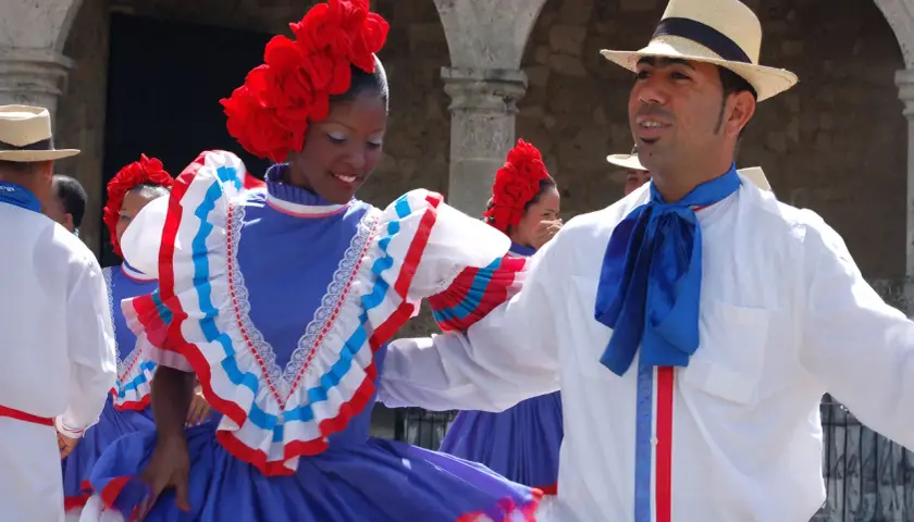 Coppia in abiti tradizionali, lei con fiori rossi sui capelli e un vestito blu con balze, lui con cappello di paglia e fiocco blu, pronti per un vivace ballo di salsa e merengue.