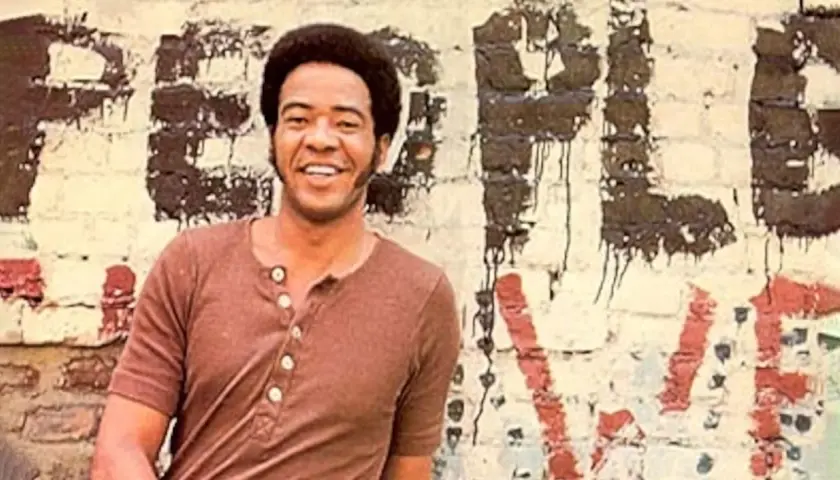 Bill Withers sorride davanti a un muro con graffiti. Anche se "ain't no sunshine", il suo sorriso illumina la scena.