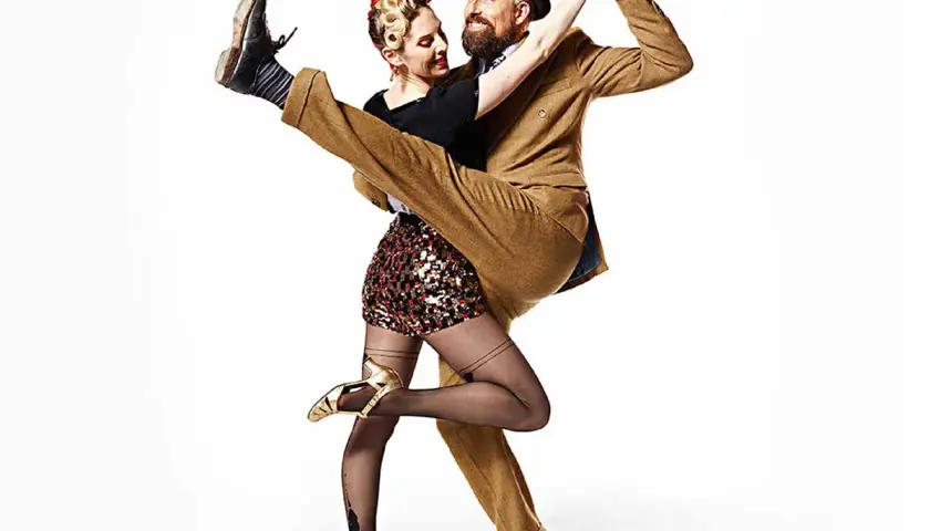 A man in a tan suit dips a woman in a sequined skirt during a Lindy Hop dance.