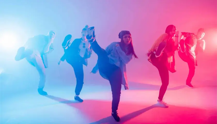 Un gruppo di ballerini esegue una coreografia hip hop con calci in aria, illuminati da luci blu e rosa.