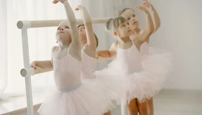 Bambine in tutù rosa praticano la danza propedeutica alla sbarra, con braccia alzate e sguardi concentrati.