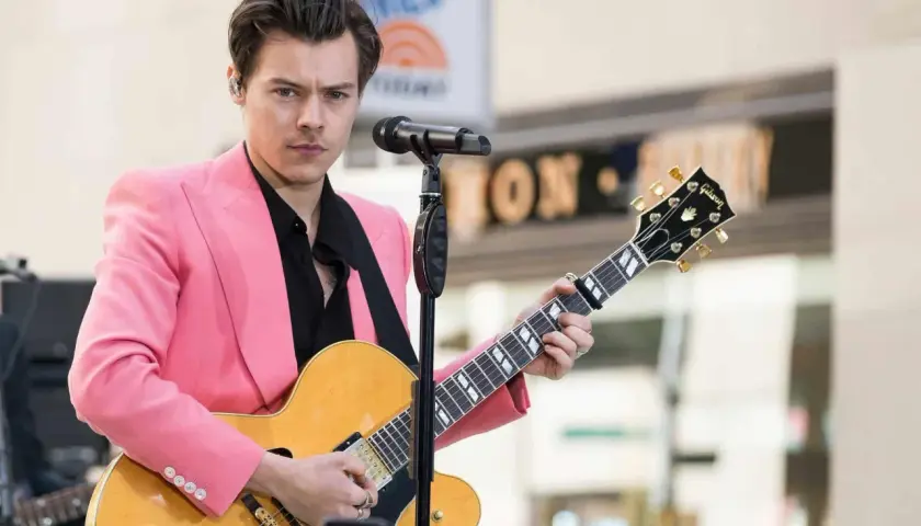 Harry Styles con giacca rosa suona la chitarra, ispirando tante canzoni da suonare con la chitarra.