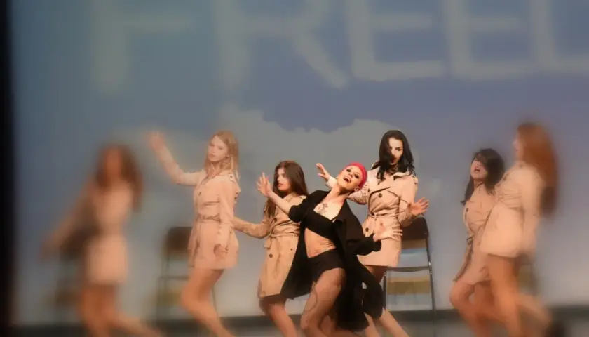 Un gruppo di ballerine in trench esegue una coreografia energica, con un'artista in primo piano che canta. Le canzoni di danza moderna creano un'atmosfera vibrante.