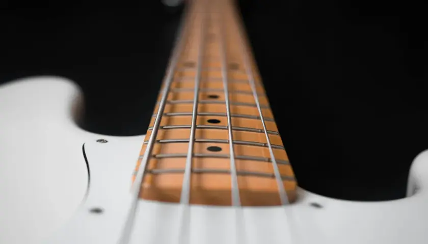 Primo piano del manico di un basso elettrico bianco, con le corde metalliche che corrono lungo la tastiera in legno chiaro.