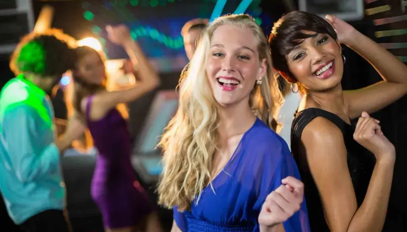 Due amiche sorridono mentre si divertono a ballare latino americano in discoteca, con luci colorate e altre persone che ballano sullo sfondo.