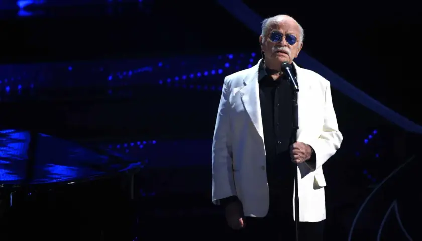 Un uomo con occhiali da sole blu canta sul palco, con un pianoforte a coda scuro e luci blu sullo sfondo. Sembra che sia lui chi ha scritto il cielo in una stanza.