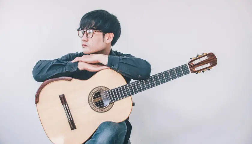 Giovane con occhiali appoggiato a una chitarra classica, pensieroso. Quanto tempo ci vuole per suonare la chitarra? Dipende dalla passione.