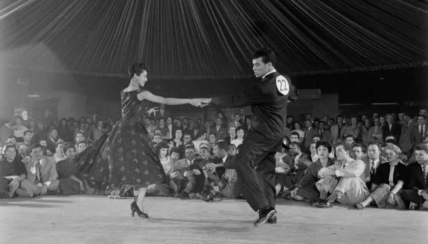Coppia in abito da sera e completo esegue una salsa danza, con pubblico seduto sullo sfondo.