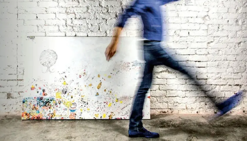 A blurred figure in blue jeans and a jacket walks past a canvas covered in colorful abstract art and doodles.