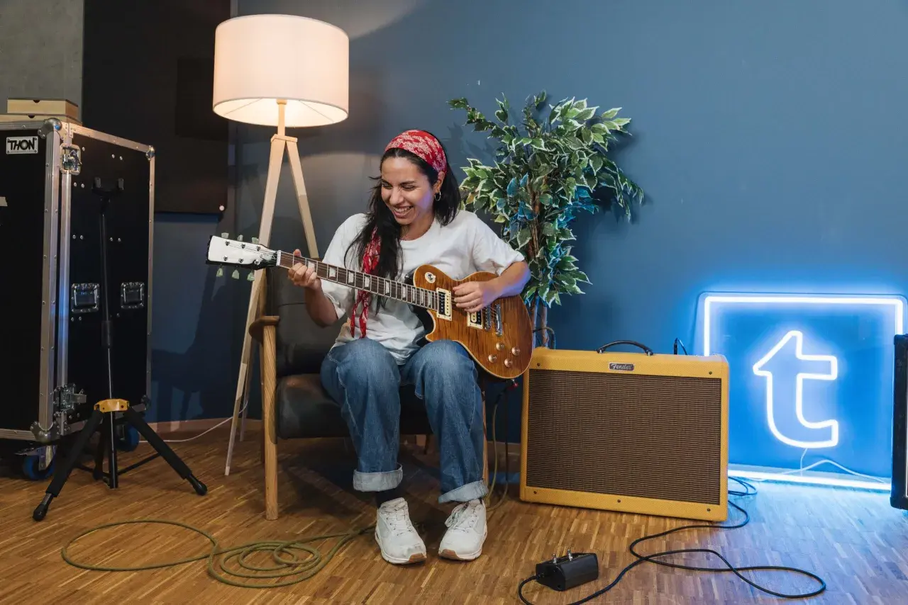 Donna sorridente suona la chitarra, con un'insegna al neon blu sullo sfondo che ricorda le "blue canzoni famose".