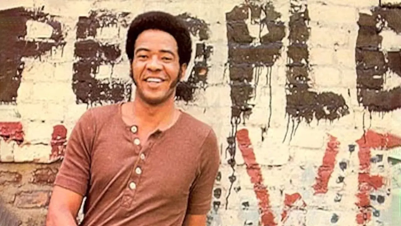 Bill Withers sorride davanti a un muro con graffiti. Anche se "ain't no sunshine", il suo sorriso illumina la scena.