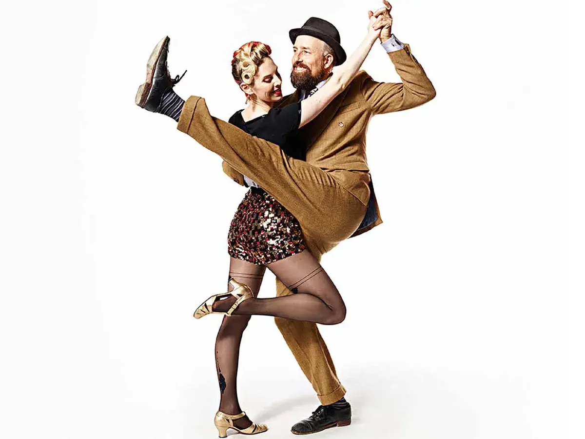 A man in a tan suit dips a woman in a sequined skirt during a Lindy Hop dance.
