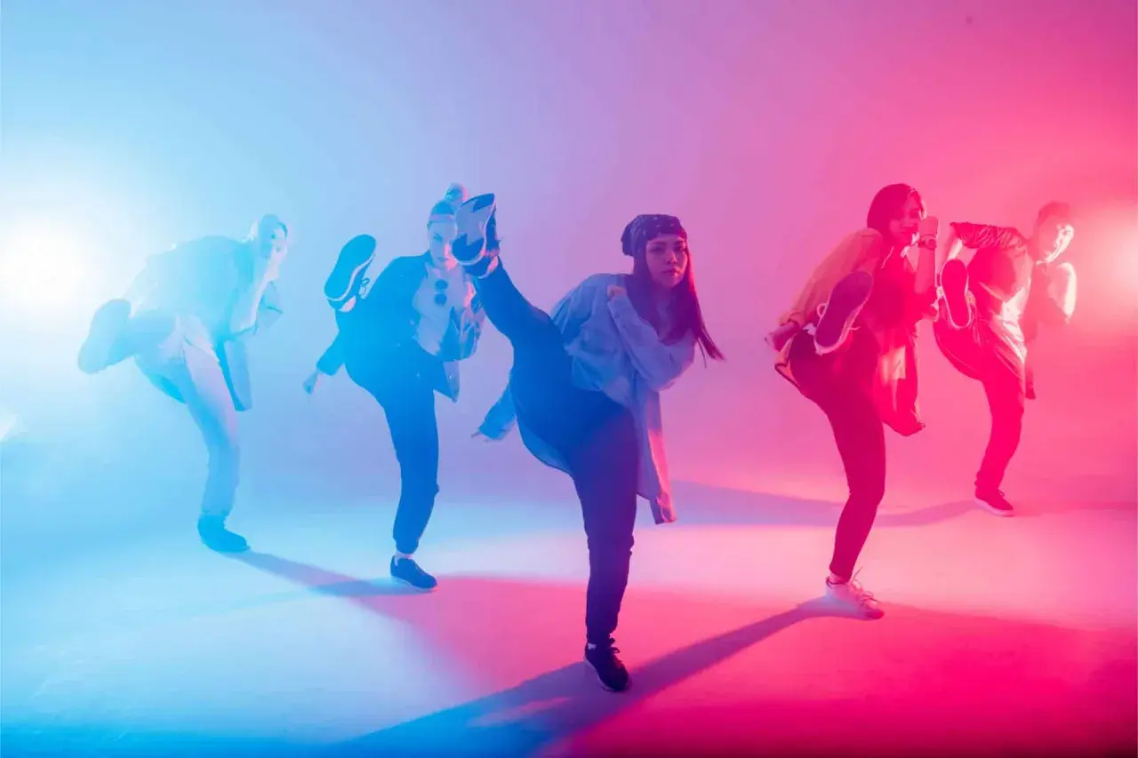 Un gruppo di ballerini esegue una coreografia hip hop con calci in aria, illuminati da luci blu e rosa.