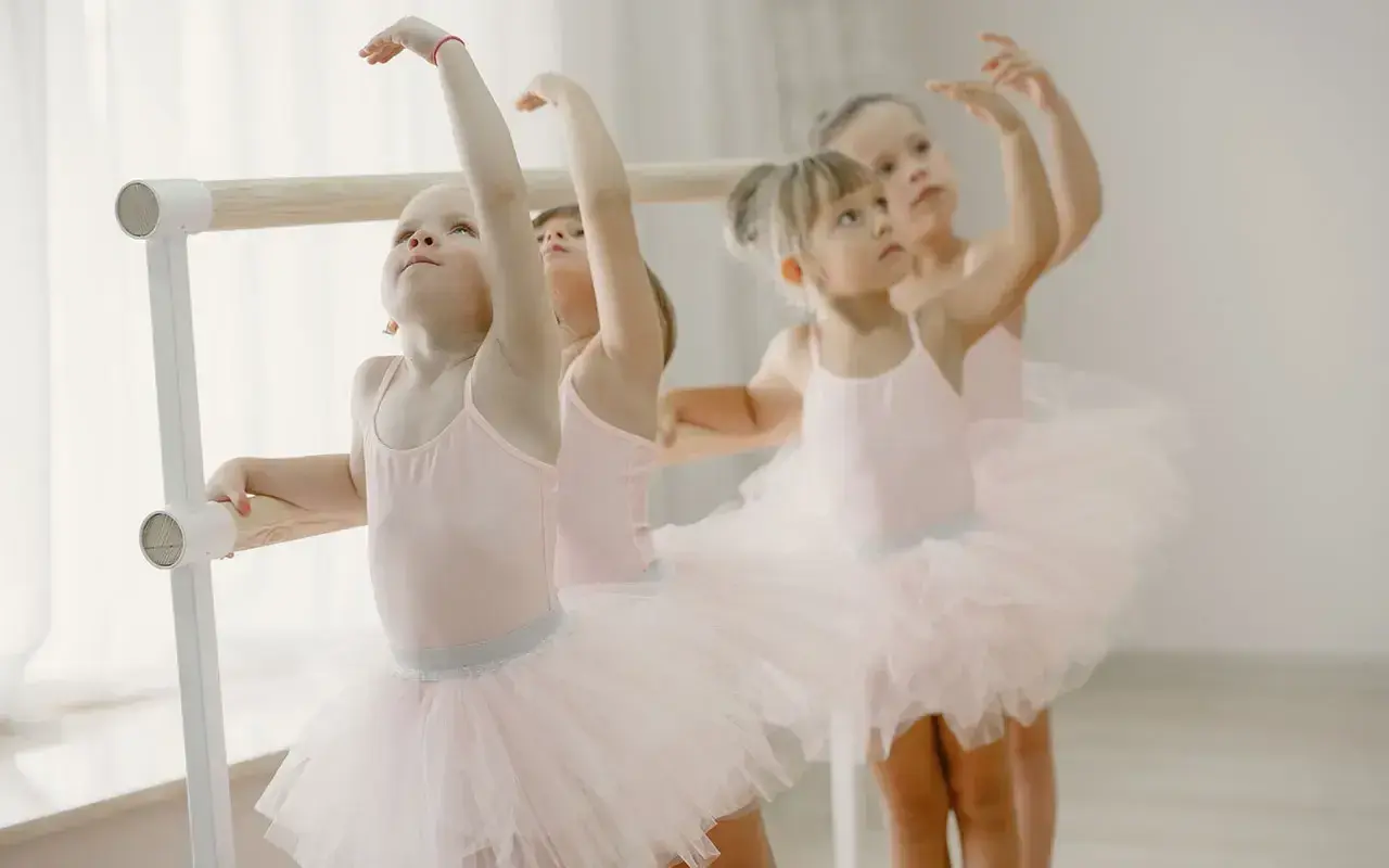 Bambine in tutù rosa praticano la danza propedeutica alla sbarra, con braccia alzate e sguardi concentrati.