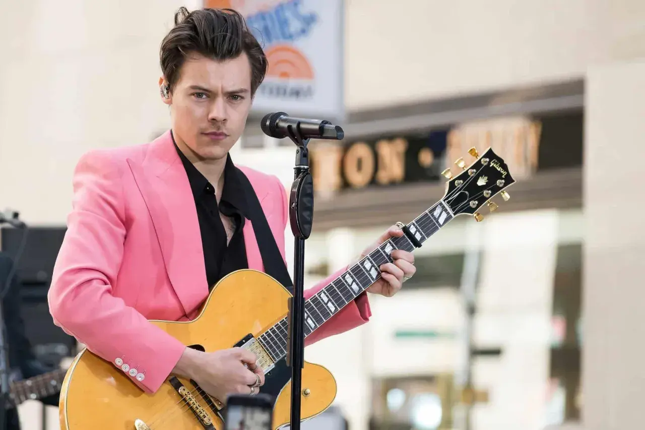 Harry Styles con giacca rosa suona la chitarra, ispirando tante canzoni da suonare con la chitarra.