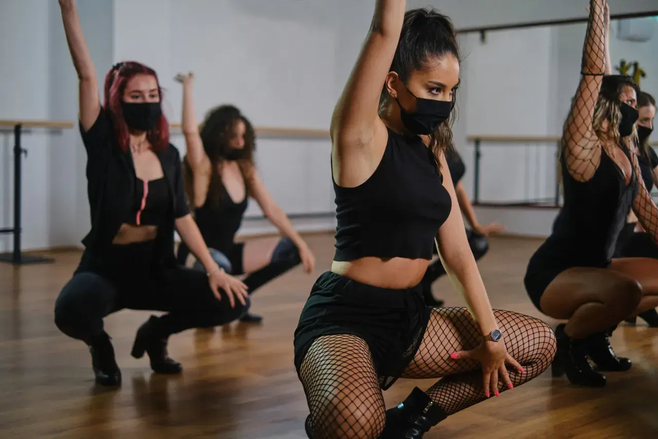 Ragazze in abiti neri e maschere eseguono una energica danza reggaeton in sala prove.