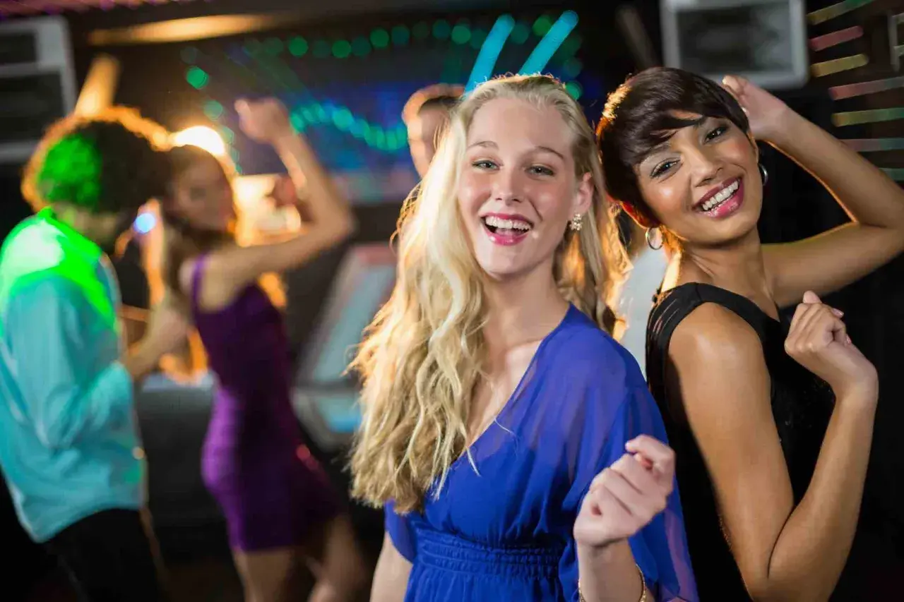 Due amiche sorridono mentre si divertono a ballare latino americano in discoteca, con luci colorate e altre persone che ballano sullo sfondo.