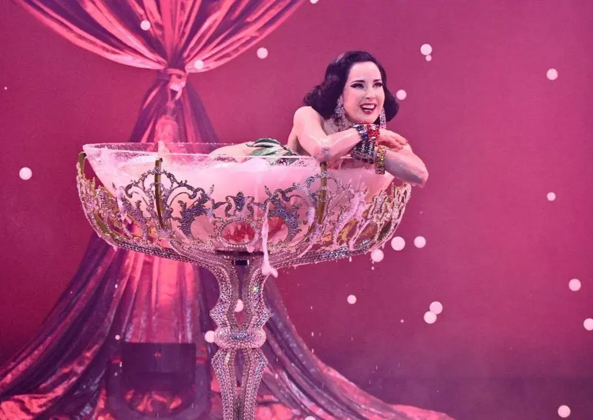 Attrice in un bicchiere gigante, un'esibizione di burlesque dance con schiuma rosa e drappeggi di seta.