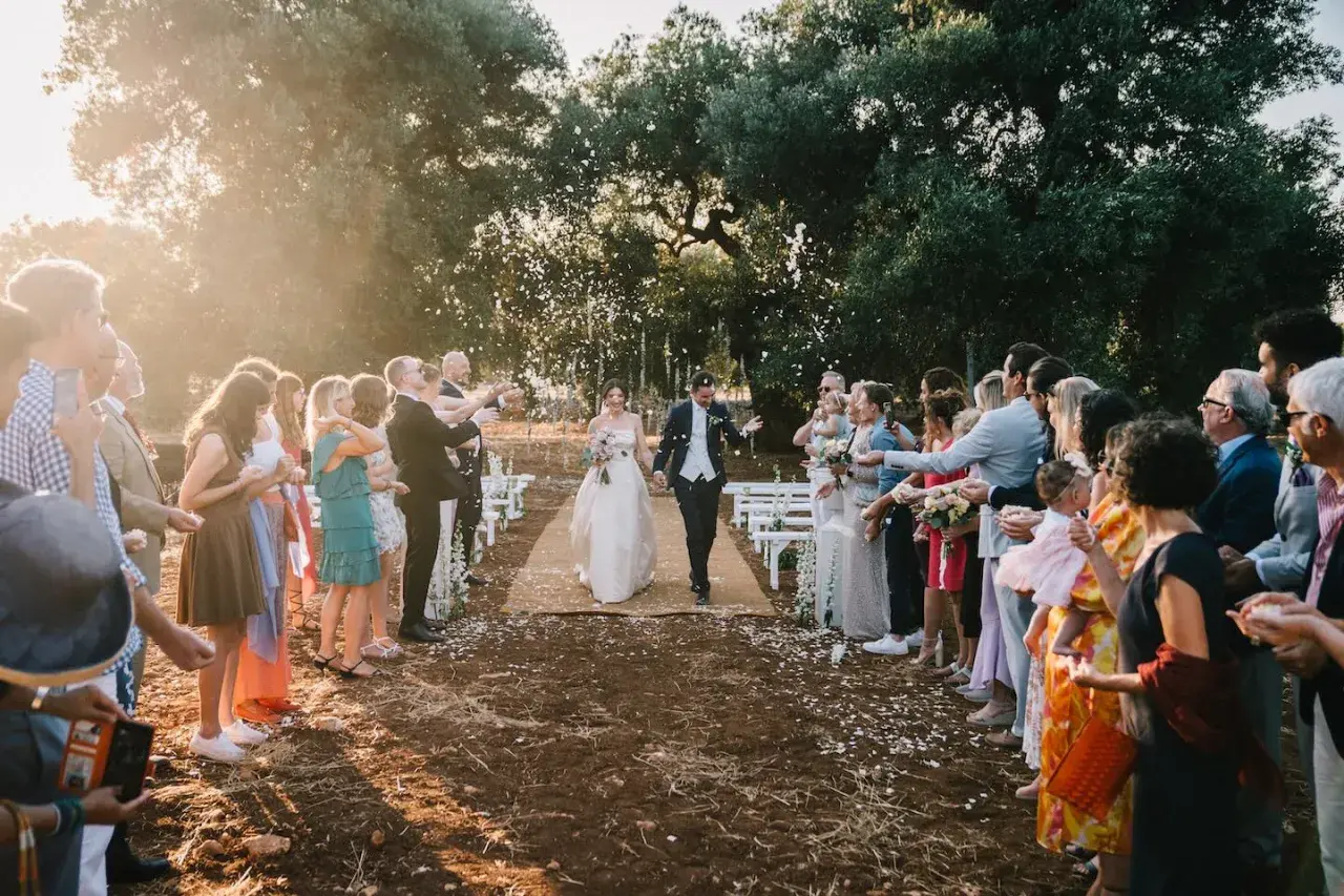 Sposi camminano su un tappeto, circondati da invitati che lanciano petali. La luce del tramonto crea un'atmosfera magica, perfetta per una canzone matrimonio.