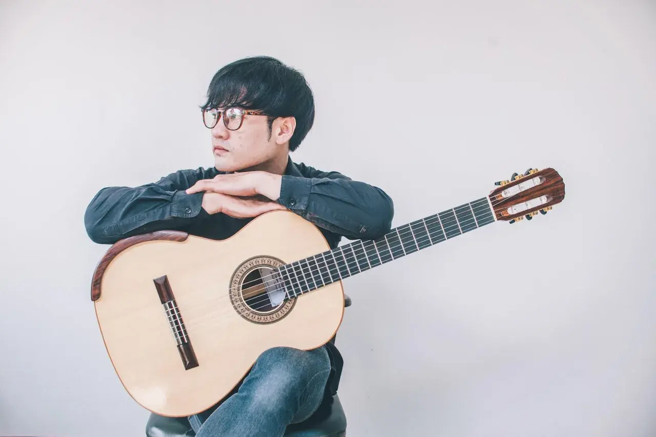 Giovane con occhiali appoggiato a una chitarra classica, pensieroso. Quanto tempo ci vuole per suonare la chitarra? Dipende dalla passione.