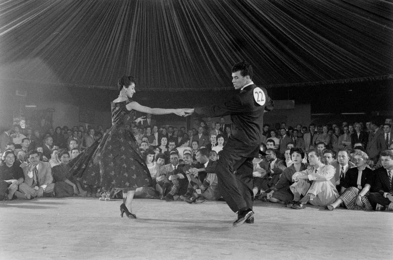 Coppia in abito da sera e completo esegue una salsa danza, con pubblico seduto sullo sfondo.