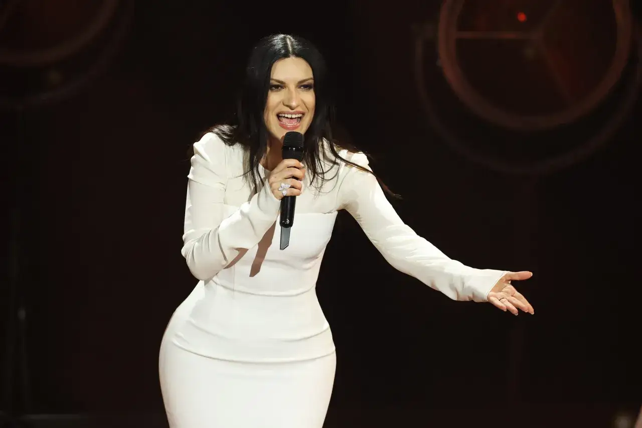 Laura Pausini, con un abito bianco attillato, canta sul palco con un microfono in mano e un sorriso radioso.
